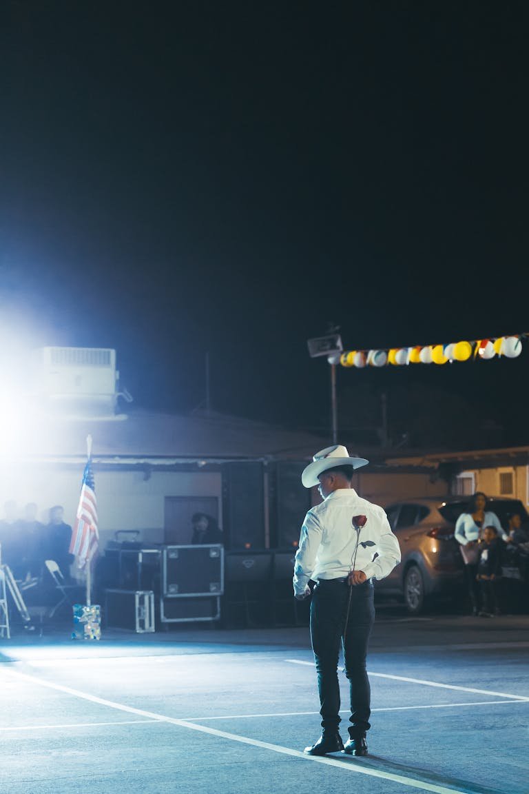 A solitary man in a cowboy hat holding a red rose in an illuminated parking lot at night.